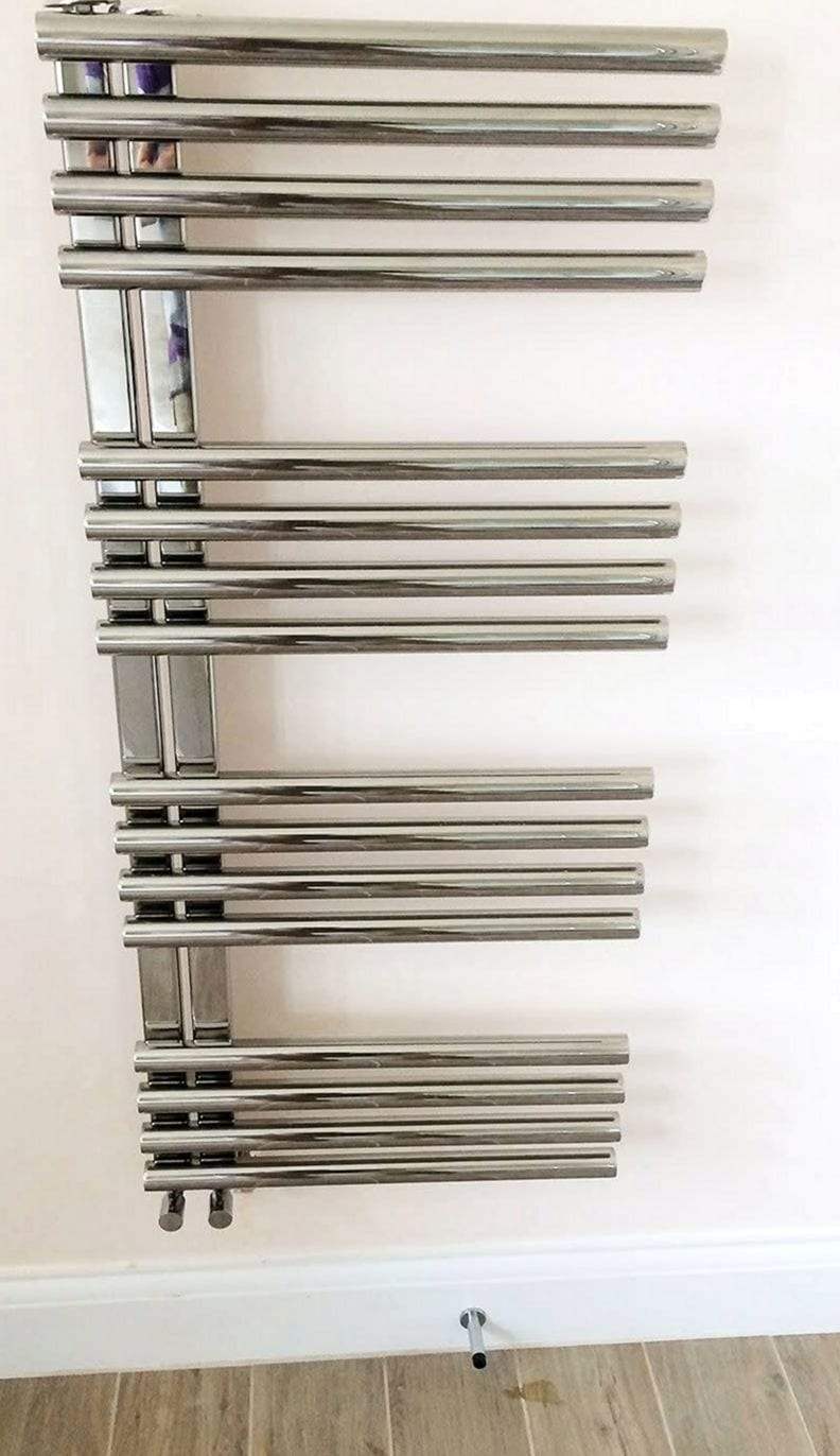 Polished chrome towel warmer with four groups of horizontal bars on a pale pink bathroom wall, its valves at the lower left and a pipe rising from the wood-look floor
