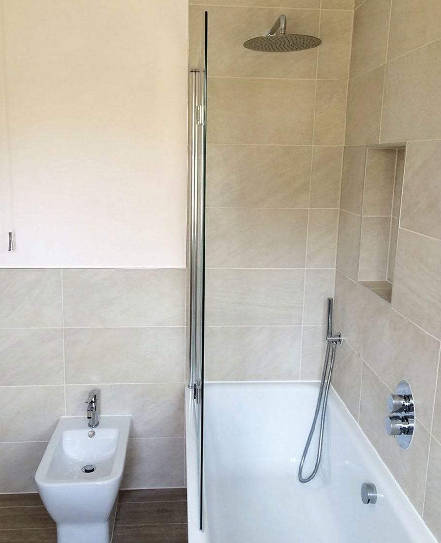 Bathtub with a hinged glass screen, a round rain shower head, a handheld shower on a slide bar, a concealed shower valve with two chrome handles and a tiled wall niche, next to a white bidet