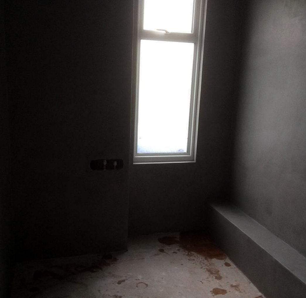 Bathroom stripped to bare plastered walls and a concrete floor, with two capped water supply stubs on the wall ready for a new fixture and a boxed-in ledge along the right wall