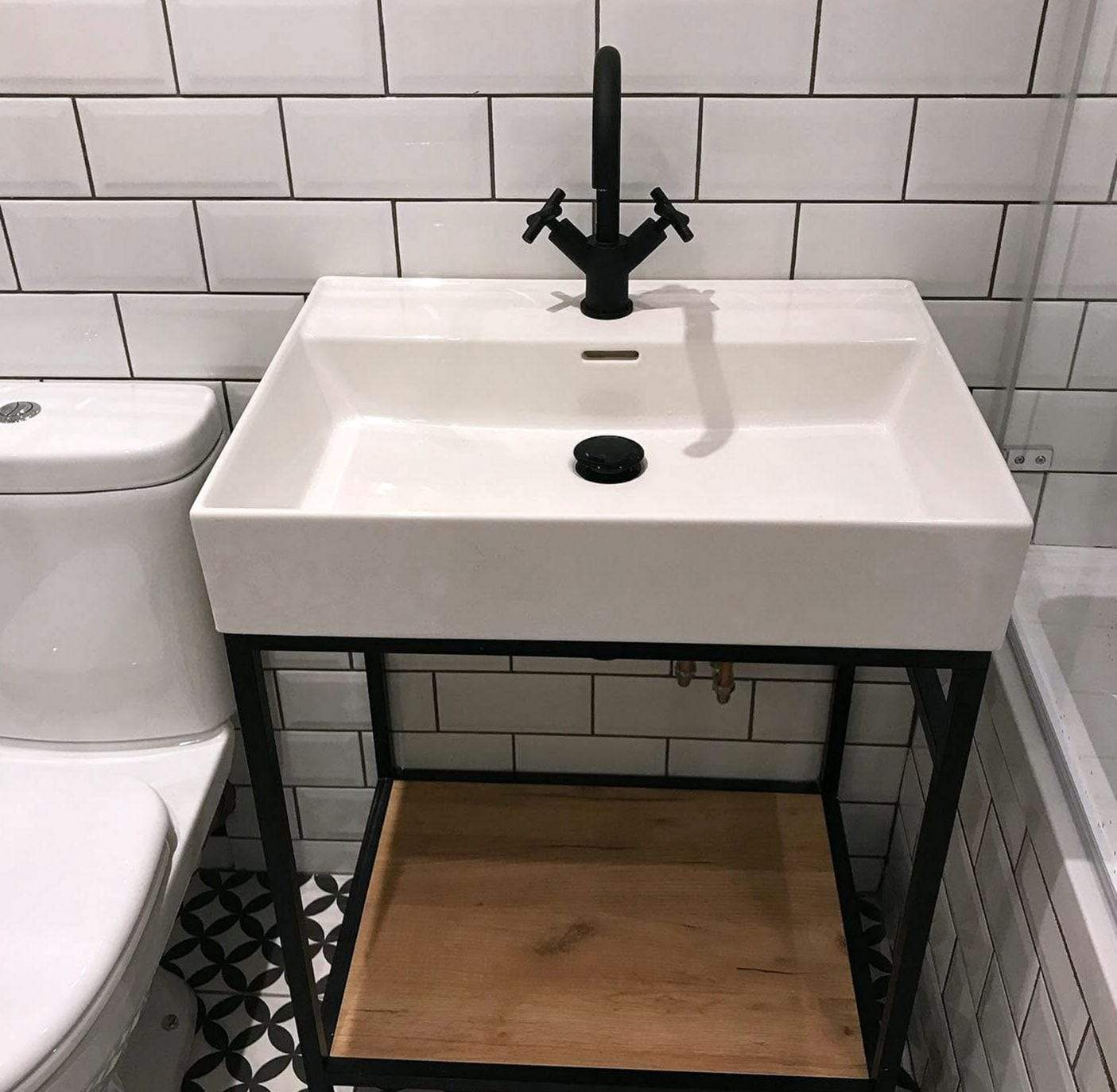 White rectangular sink on a black metal washstand with a wood shelf, a matte black faucet with cross handles, white subway tile with dark grout and copper supply pipes behind the stand