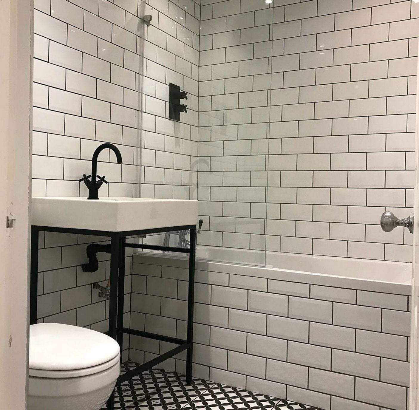 Small bathroom in white subway tile with dark grout: a sink on a black metal stand, a black shower valve above the tub, a glass tub screen, a toilet and black-and-white patterned floor tile