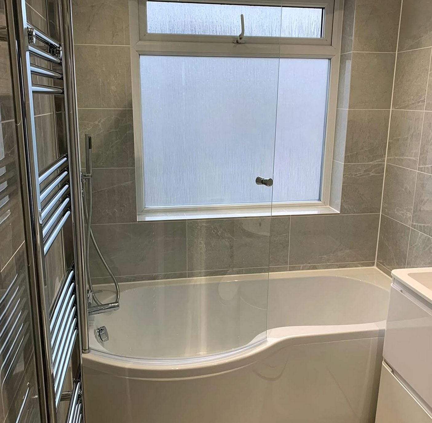 Curved shower tub with a glass screen, a chrome handheld shower on a slide bar, gray marble-look wall tile, a frosted window and a tall chrome towel warmer