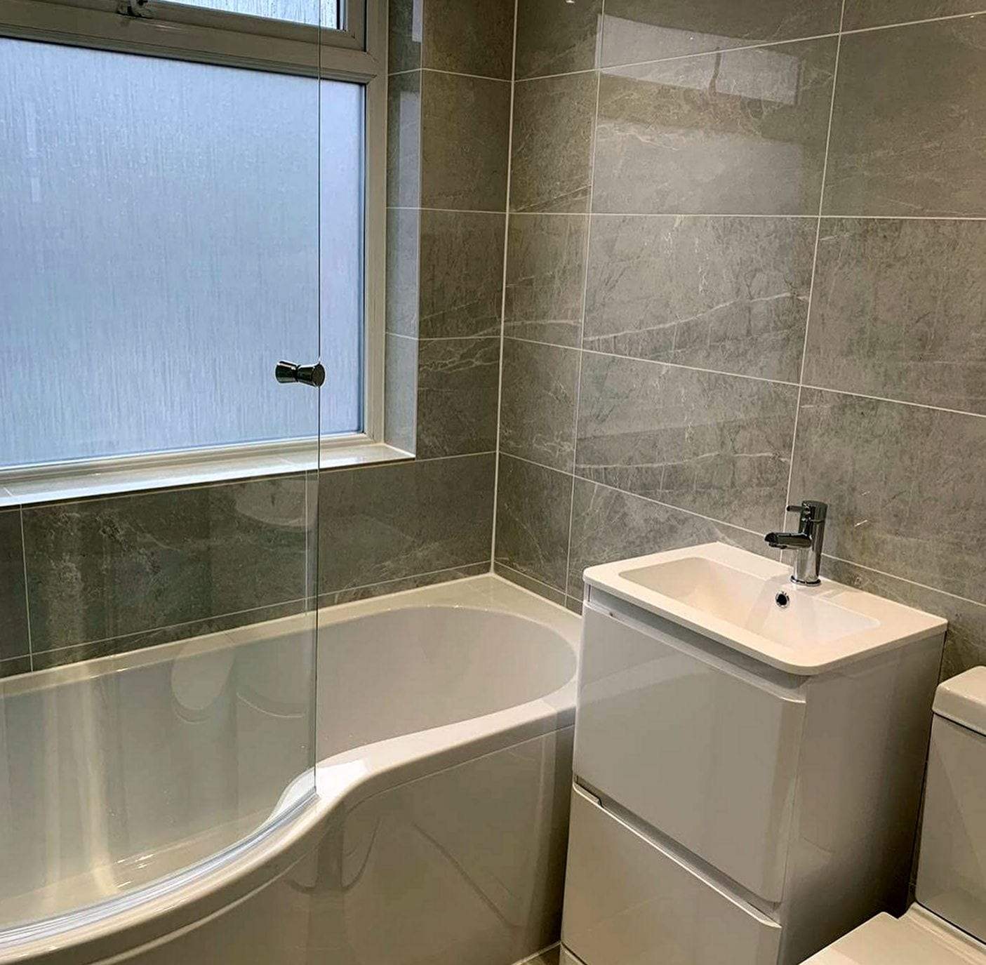 Gray-tiled bathroom with a curved shower tub and glass screen beside a white gloss vanity with a built-in sink and a chrome single-handle faucet