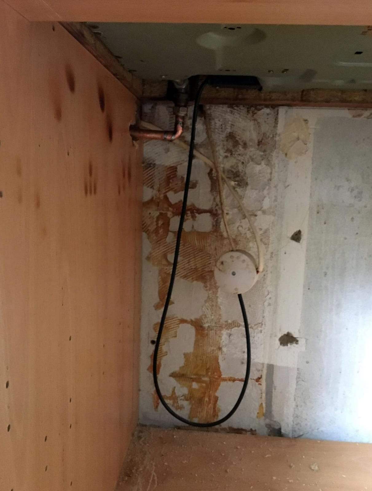 Inside an empty kitchen base cabinet with the back panel removed: a copper pipe with an elbow and a fitting under the counter, a black cable looping down and old adhesive marks on the bare wall