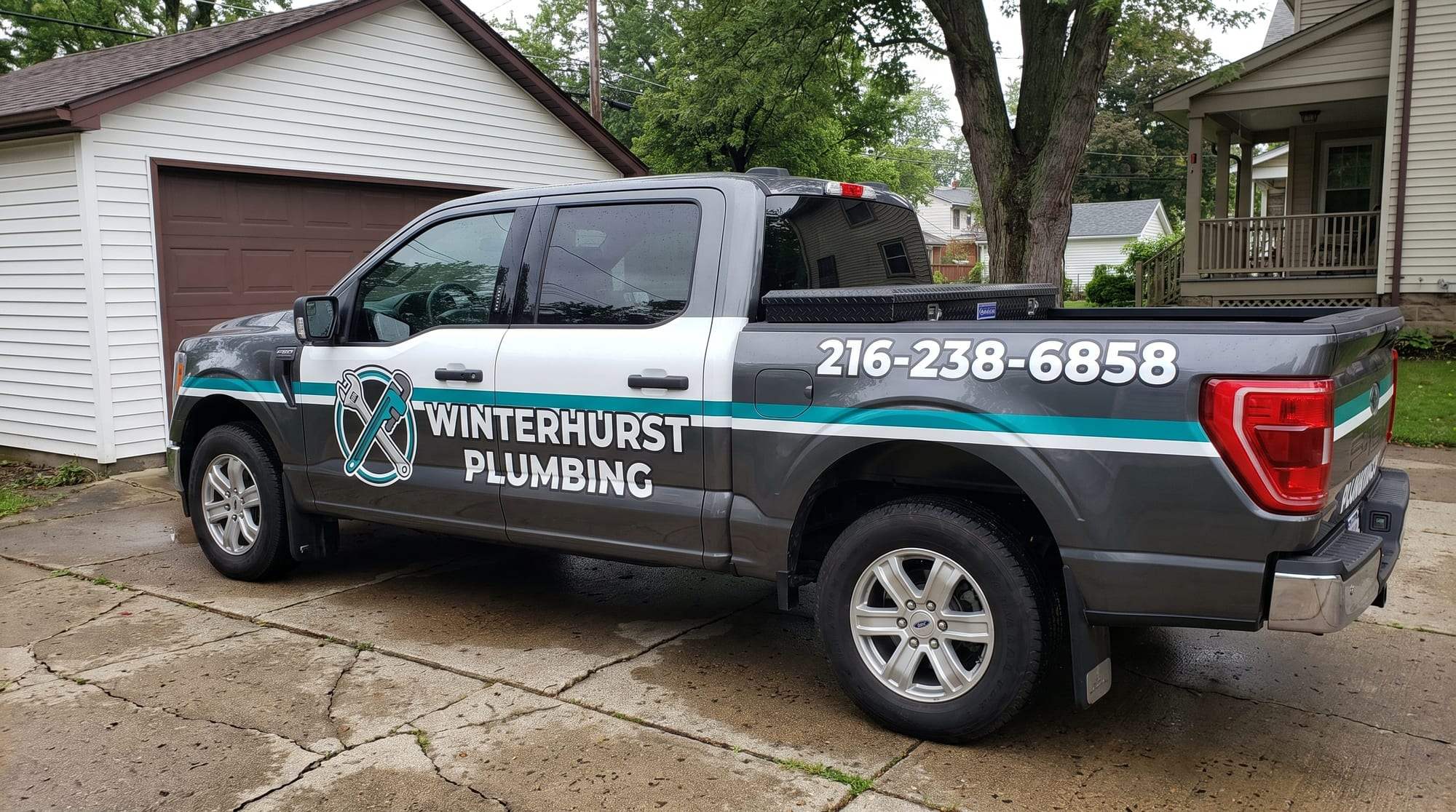 Dark gray Winterhurst Plumbing pickup truck with a white and teal stripe, the crossed-wrench logo on the rear door and the phone number 216-238-6858 on the bed, parked on a concrete driveway beside a white garage and a house with a front porch