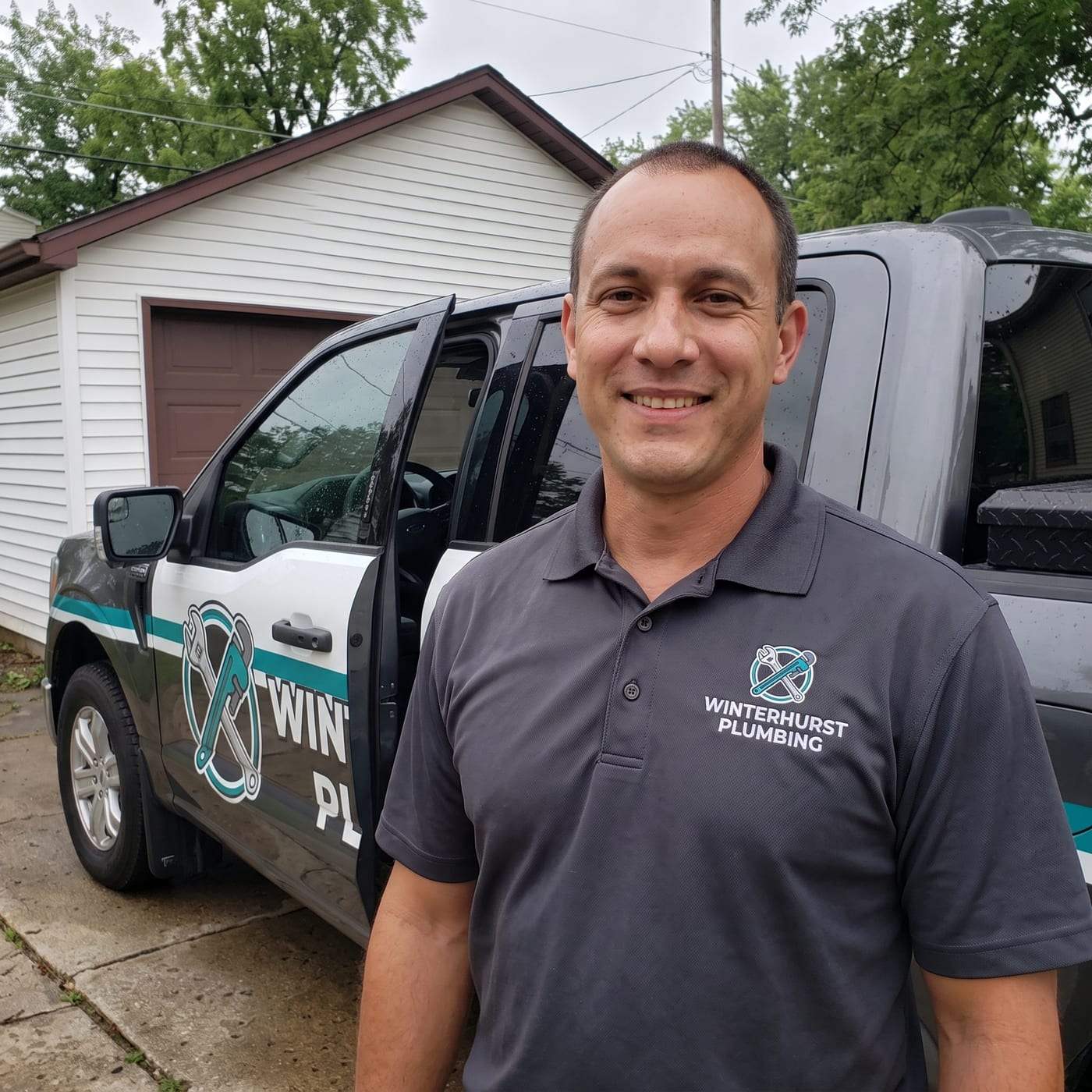 Jonas Lewis, manager of Winterhurst Plumbing, smiling in a dark gray polo shirt with the company logo, standing beside the lettered service truck in a driveway